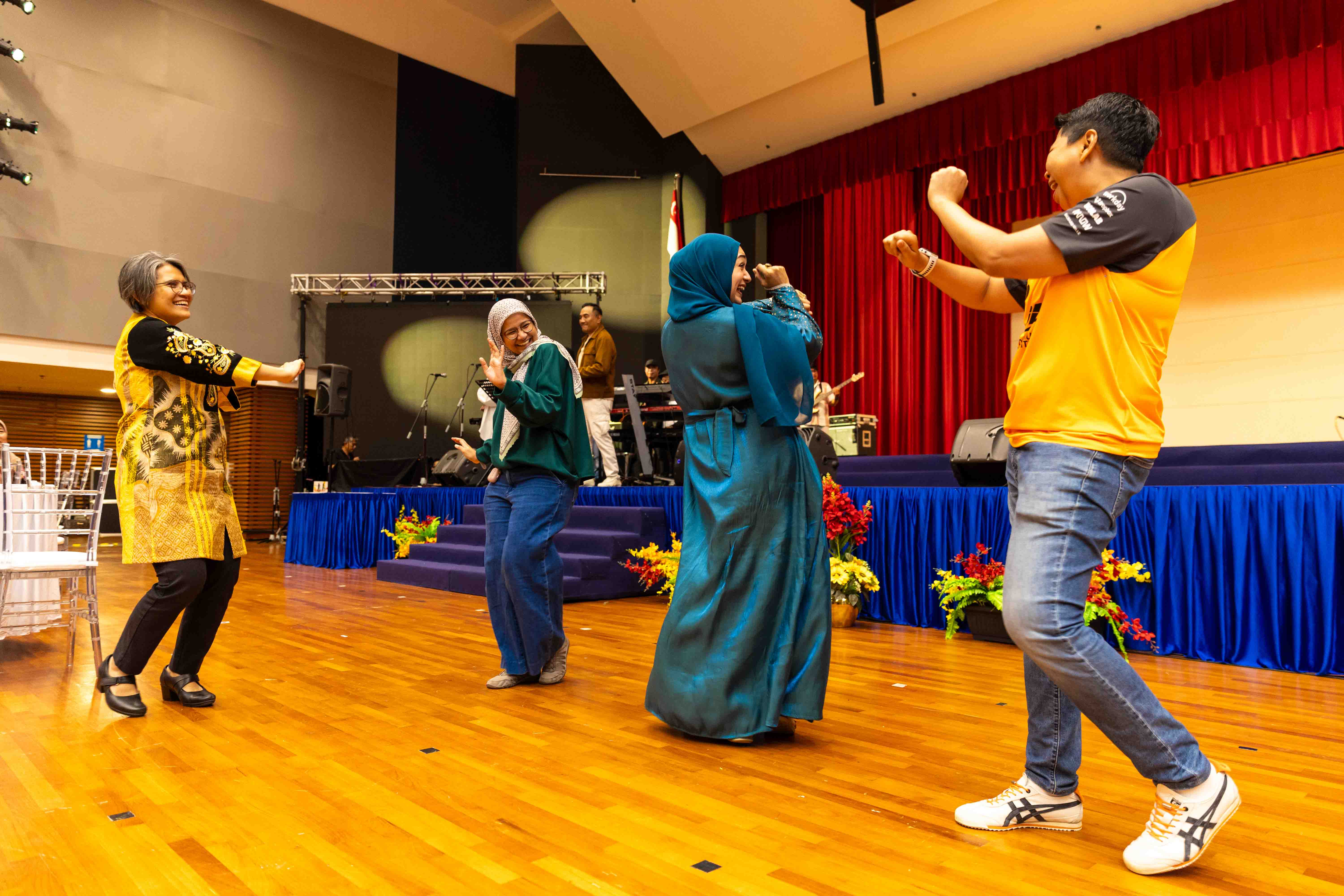 Four participants dance together on the hall floor in front of the stage.